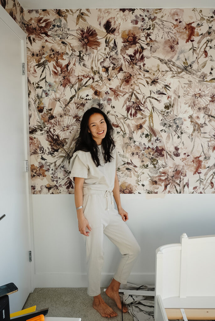 learning about installing glue-based wallpaper with wallpaper on half of a wall. the wallpaper is a beautiful floral print with creams, magentas, and blue hues. Samantha Potter is standing in front of the wallpaper.