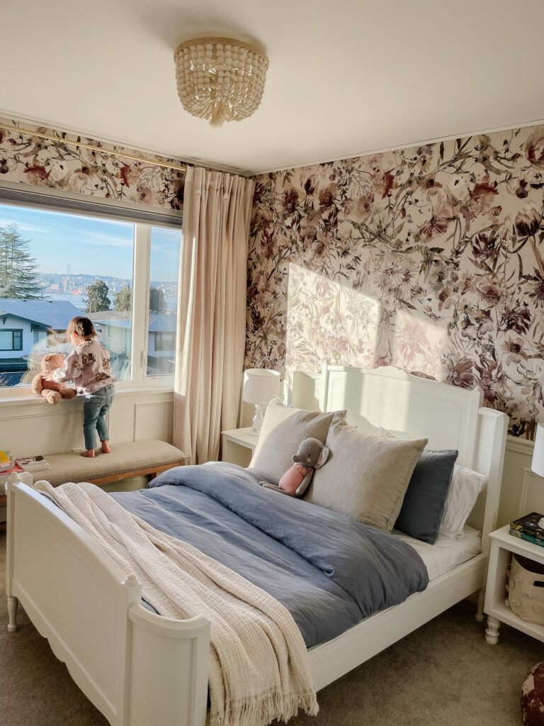 Sloane sitting in her big girl room with wallpaper on the walls, chair railing, velvet ivory curtains, a blue comforter