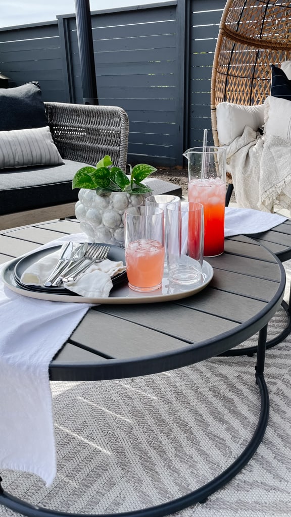 Outdoor patio oasis set up with club chairs, an inviting table setting with a red drink and cups, and green plants.