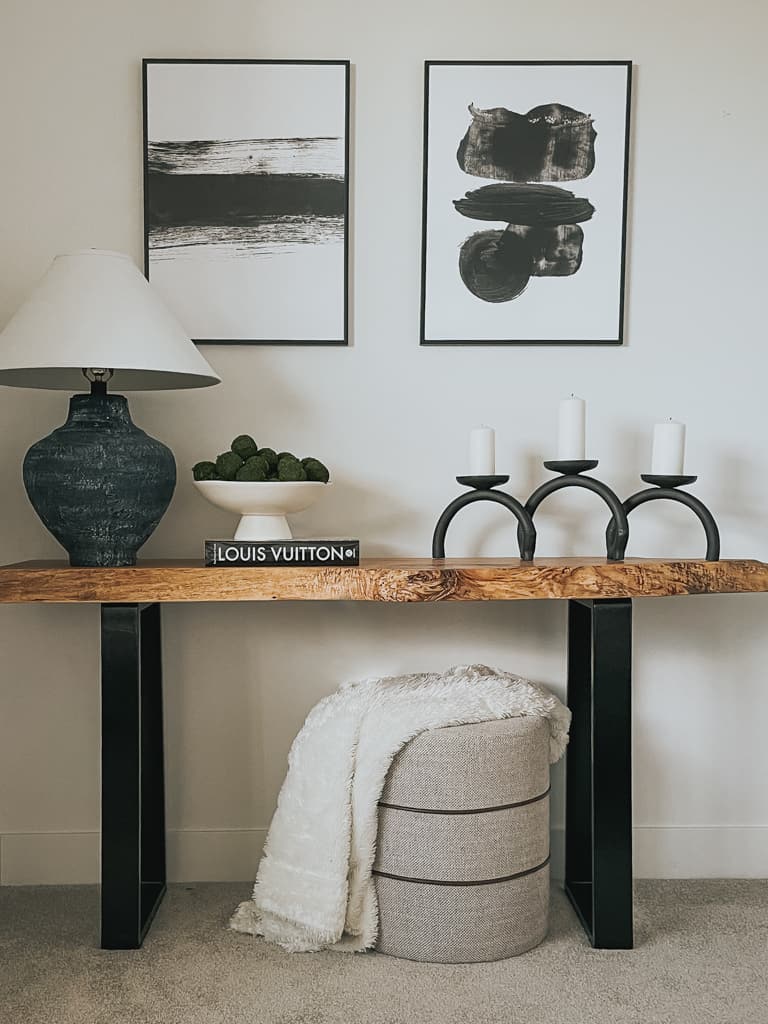 A blanket styled on top of a smaller round ottoman underneath a console table that has a lamp, canadles, and book on it.