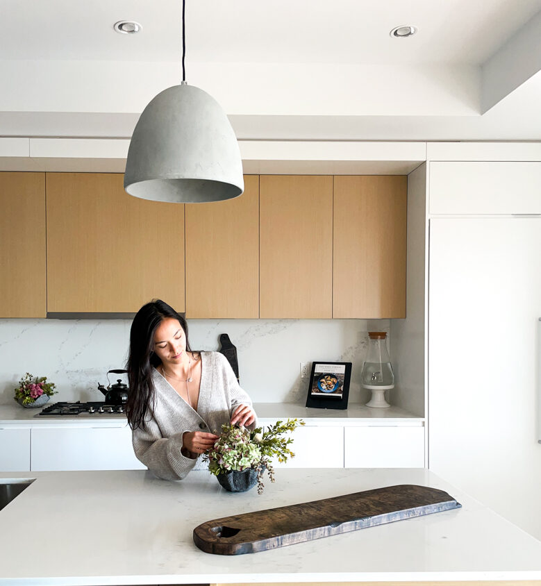 Samantha standing in her kitchen arranging her DIY gifts - including adding florals to a DIY concrete bowl and a DIY charcuterie board in front of her.