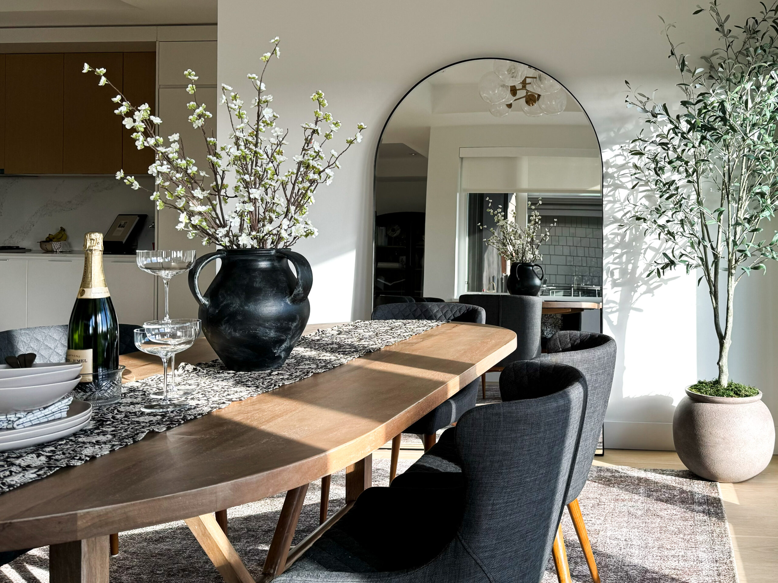 Dining room shot of the dining table with plates bowls champagne flutes florals and a runner on it. Dining room shot of the dining table with plates bowls champagne flutes florals and a runner on it. Oversized mirror in the background and olive tree