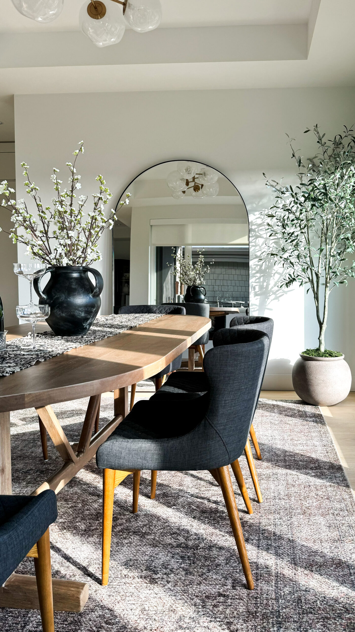 Dining room shot of the dining table with plates bowls champagne flutes florals and a runner on it. Dining room shot of the dining table with plates bowls champagne flutes florals and a runner on it. Oversized arched mirror in the background. And an olive tree. Faux spring stems in a vase on the table with cherry blossoms