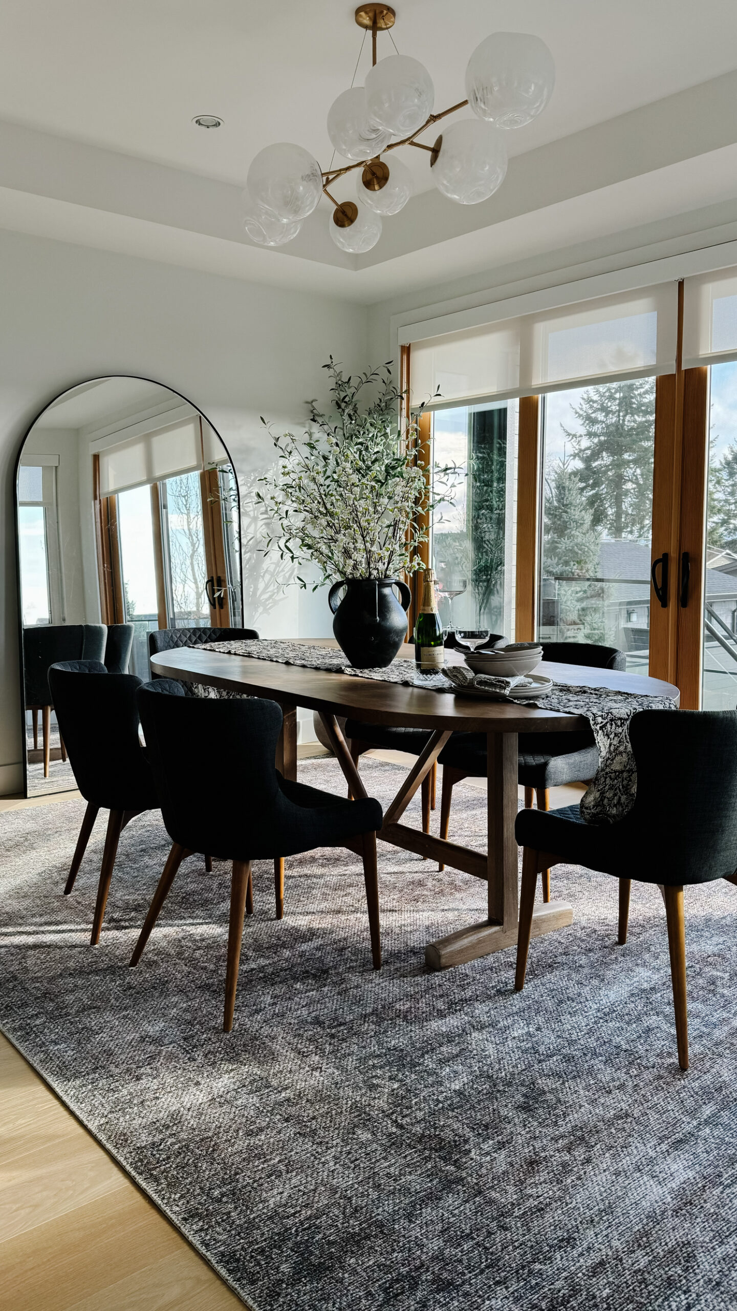 Dining room shot of the dining table with plates bowls champagne flutes florals and a runner on it. Dining room shot of the dining table with plates bowls champagne flutes florals and a runner on it. Oversized mirror in the background and olive tree